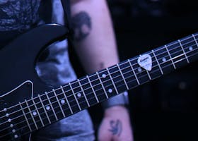 Detailed close-up of a black electric guitar in a moody setting, featuring tattooed arm.